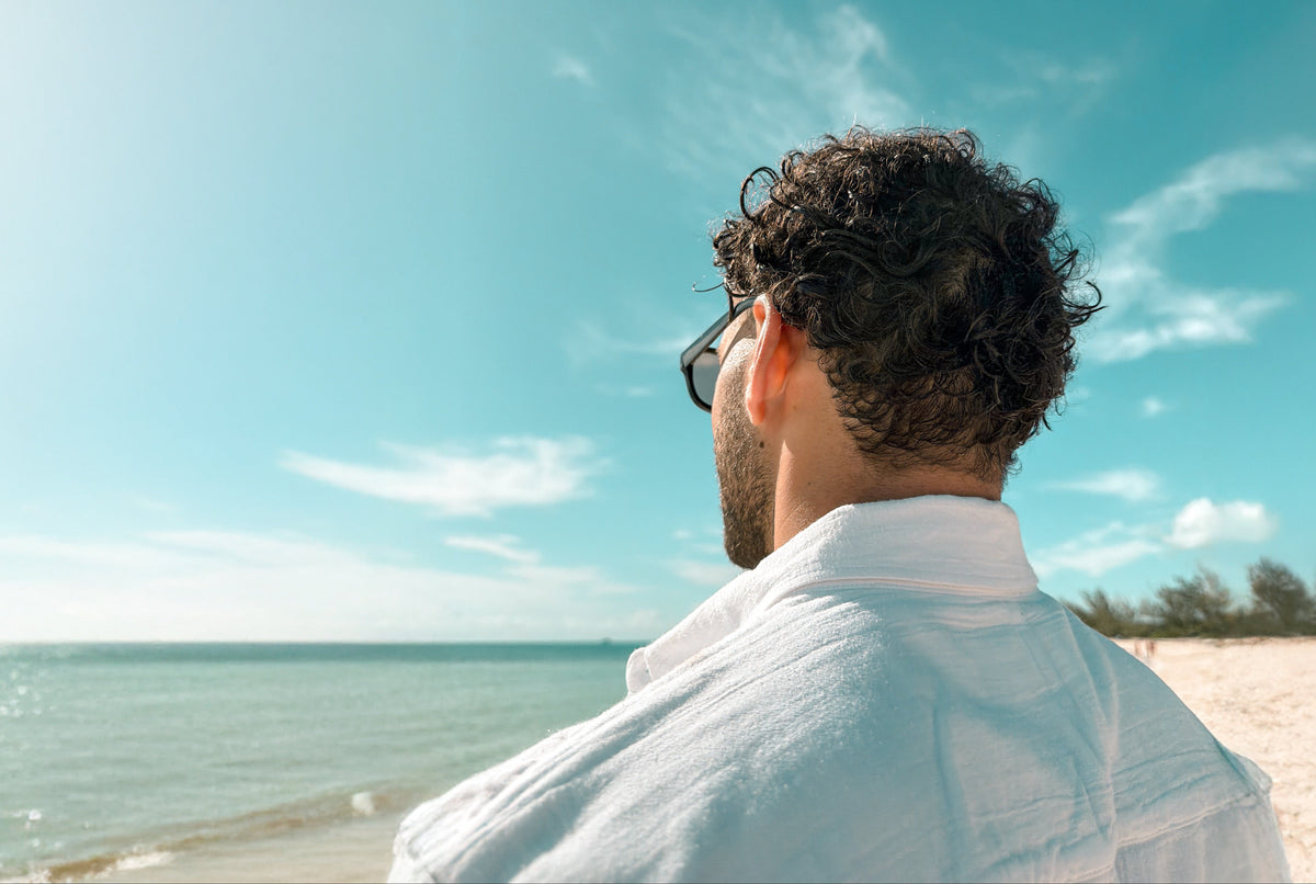 A man with curly dark hair and glasses, wearing a white shirt, is seen from behind looking out at a sunny beach and ocean under a bright blue sky.